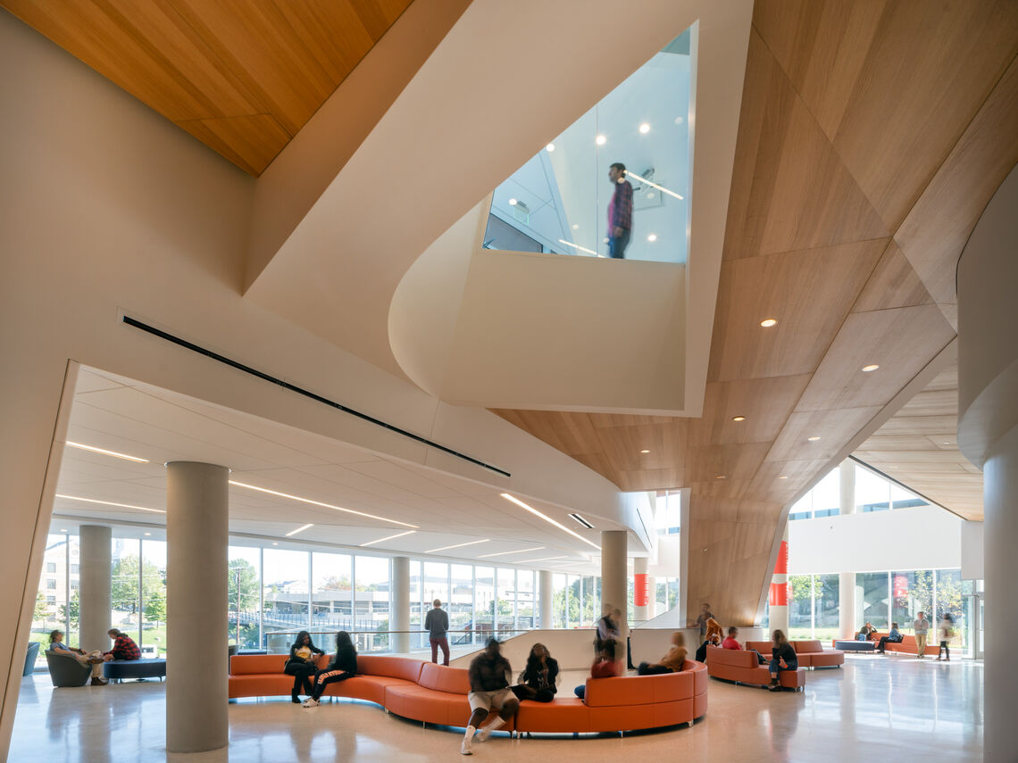 Inside, the building features reception areas and service desks organized along a series of flowing, multi-story lounges with seating, study, and collaboration spaces. A monumental stair with gently curved landings and a continuous wood soffit forms the connective tissue between the three levels of public lobbies and lounges, anchored by a student services counter.